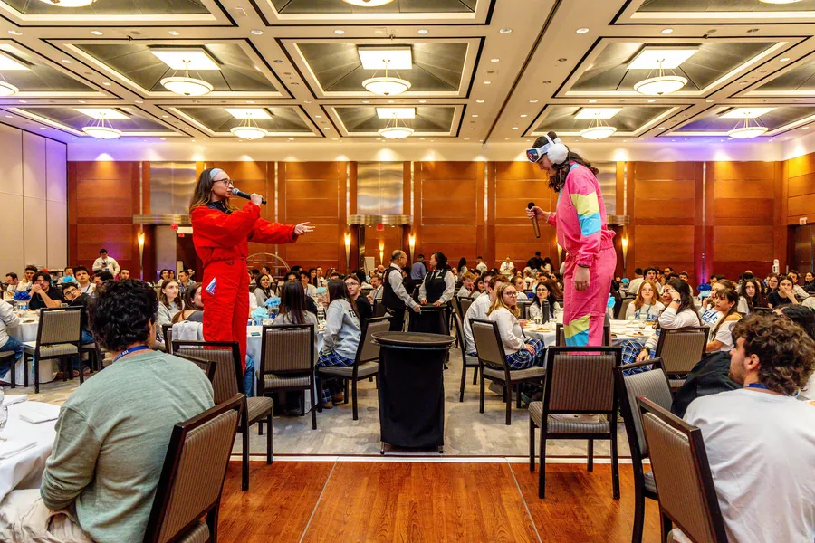 A speaker in pink addressing the full room at a plenary.